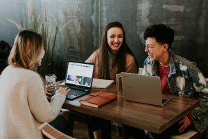 A foto traz três jovens, sorridentes, trabalhando juntos em uma pequena mesa com seus notebooks. Imagem ilustrativa do texto quais os segmentos de franquias mais rentáveis.