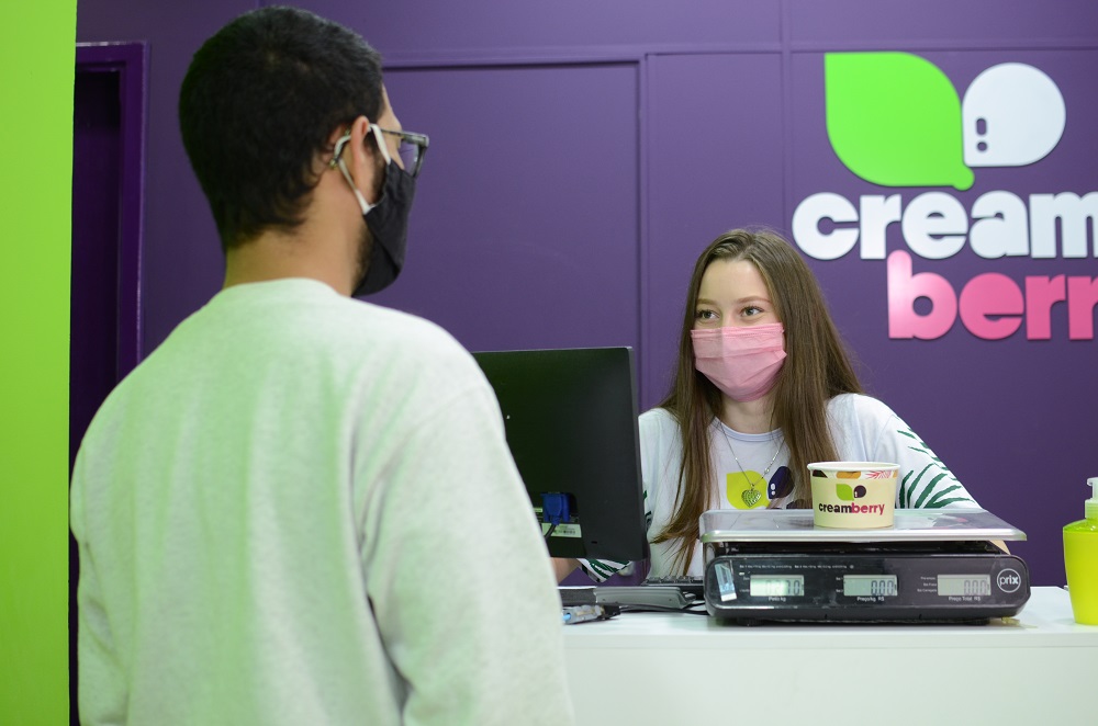 moça atendendo cliente no balcão da loja cream berry. ambos usam máscara de proteção contra a covid-19. imagem ilustrativa texto setor de alimentação