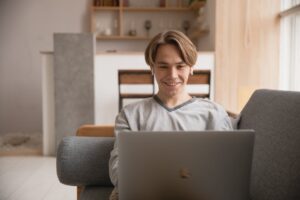 Homem sorridente, usando blusa cinza, enquanto trabalha no notebook. Ilustração do texto sobre tendências para empreender.