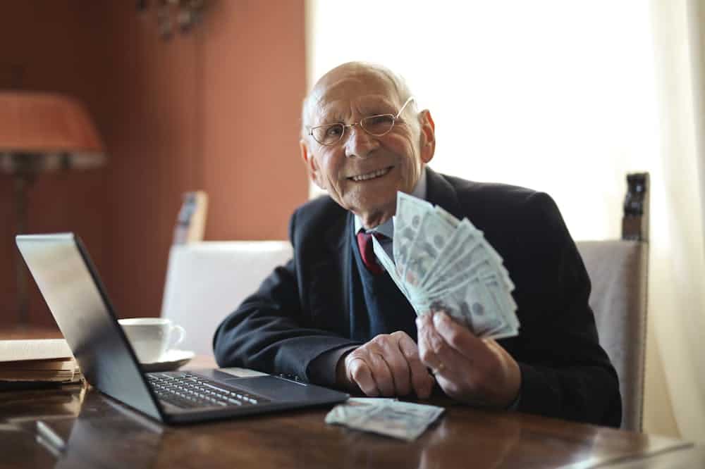 Homem idoso sorridente, usando terno cinza, segurando notas de dinheiro, enquanto está em seu escritório. Ilustração do texto sobre investidor anjo.