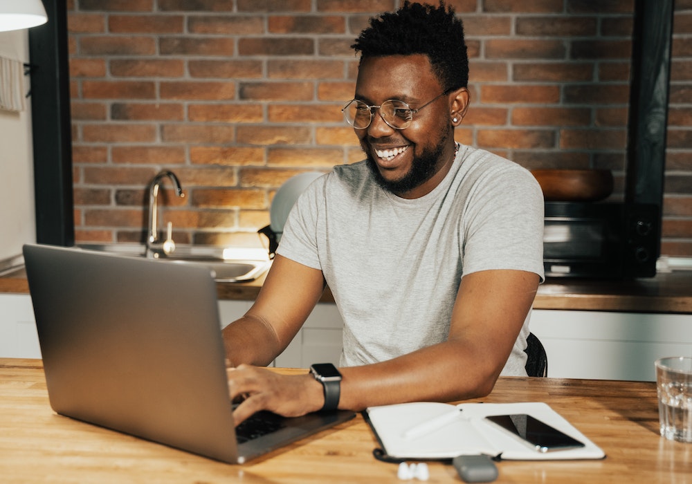 Homem de camisa cinza sorri enquanto digita no notebook. Imagem ilustrativa texto empreender em casa.