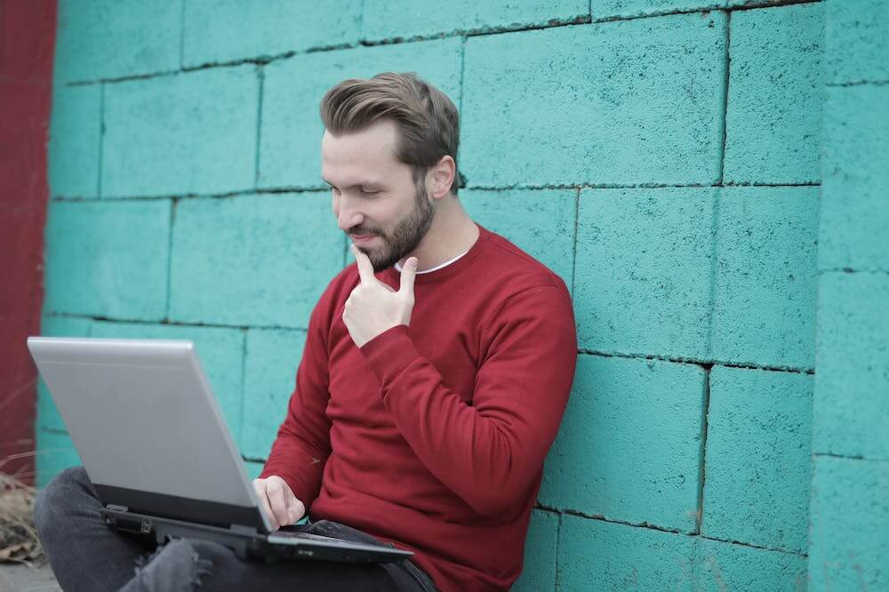 Homem pensativo, usando blusa na cor rubra, escorado em muro verde, enquanto trabalha no computador. Ilustração do texto sobre royalties, o que é.
