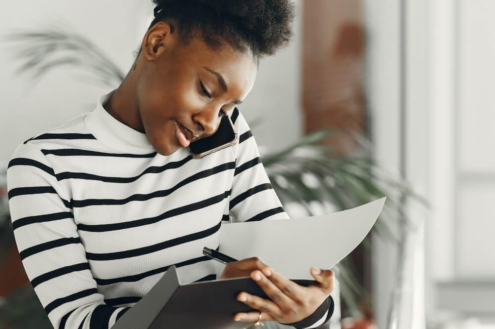 Mulher com leve sorriso, usando blusa com mangas longas listrada em preto e branco, fazendo anotações enquanto está ao telefone. Ilustração do texto sobre franqueados de sucesso.