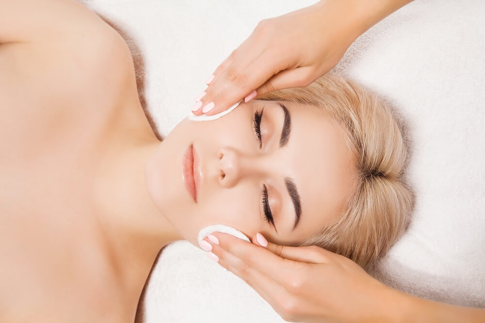 A foto traz uma mulher branca, loira de olhos fechados enquanto as mãos de uma esteticista passa um algodão pelo seu rosto. Imagem ilustrativa do texto: franquia beleza e centro de estética.