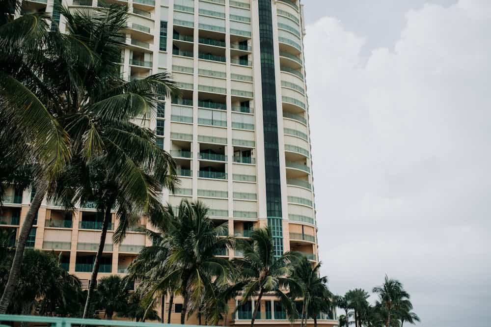 Prédio com janelas de vidro rodeado com árvores com folhas verdes. Ilustração do texto sobre franquia de administração de condomínios.