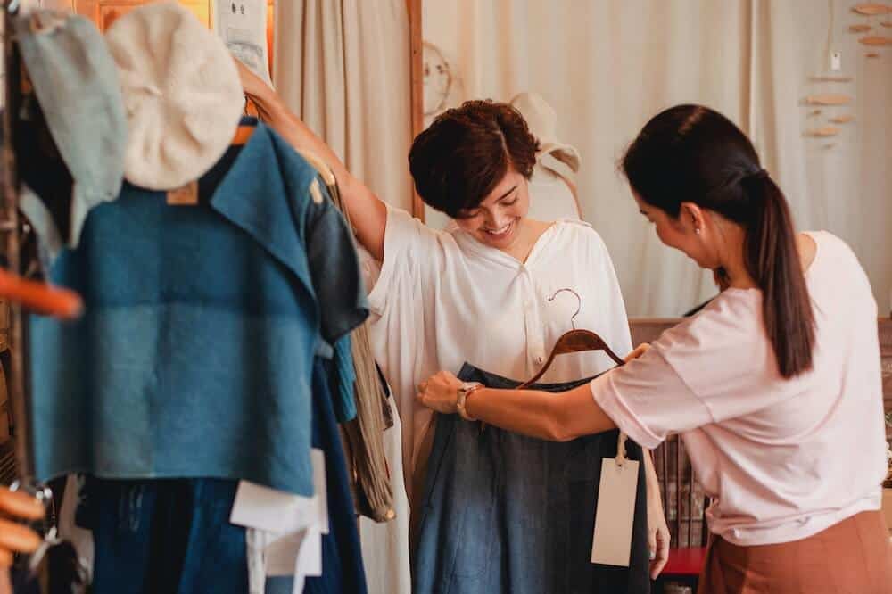 Mulheres usando camisa branca e provando roupas em uma loja. Ilustração do texto sobre franquia de roupas femininas baratas.