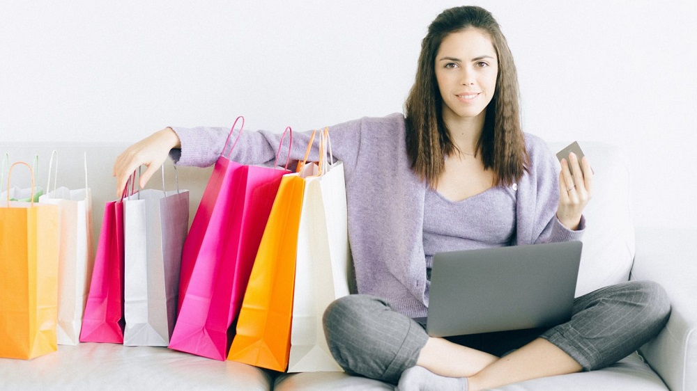 Mulher sorridente, sentada no chão, enquanto segura cartão de crédito e sacolas coloridas: rosa, amarelo e branca. Ilustração do texto sobre ticket médio.