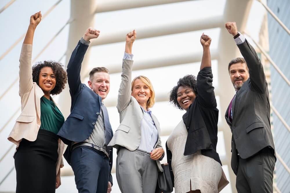 A foto traz um grupo com cinco pessoas com idades e etnias diversas, usando roupa social, sorridentes, com um dos braços para o alto. Imagem ilustrativa do texto investir em franquias vale a pena.
