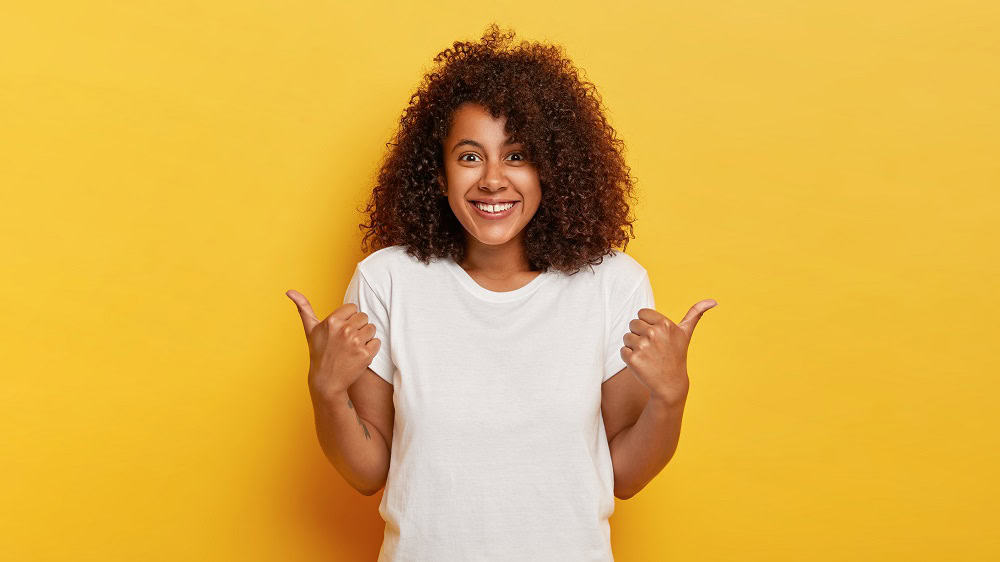 Mulher com os cabelos cacheados, usando blusa branca, sorri e faz sinal de joinha em frente a um fundo amarelo. Ilustração do texto sobre como começar a empreender.
