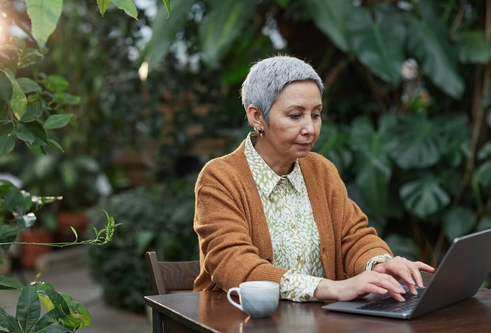 A foto traz uma mulher madura, com cabelos brancos, sentadas em uma mesa na área externa de uma casa, trabalhando em seu notebook, Imagem ilustrativa do texto negócios home office lucrativos veja opções de investimento para quem quer trabalhar em casa.