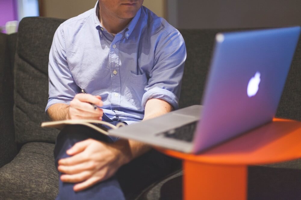 Imagem de um homem sentando ao lado de um computador com um caderno no colo fazendo anotações. Imagem ilustrativa texto empreender do zero em 2023.