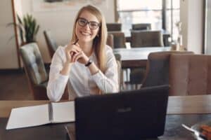 Mulher sorridente, usando blusa social branca, sentada em uma mesa de escritório, em frente a um computador. Ilustração do texto sobre franquias baratas e lucrativas.