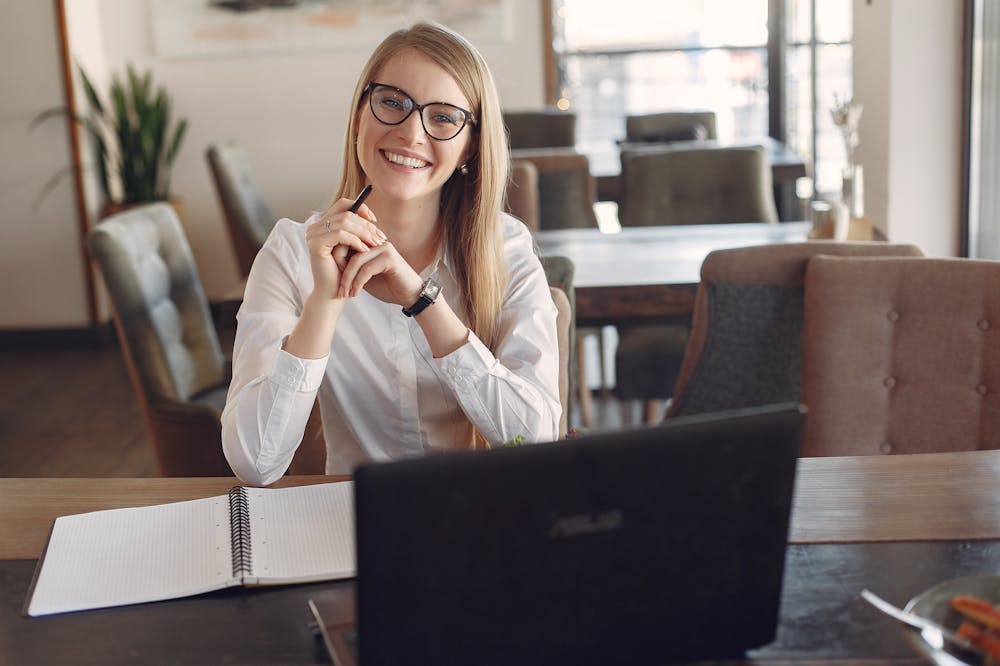 Mulher sorridente, usando blusa social branca, sentada em uma mesa de escritório, em frente a um computador. Ilustração do texto sobre franquias baratas e lucrativas.