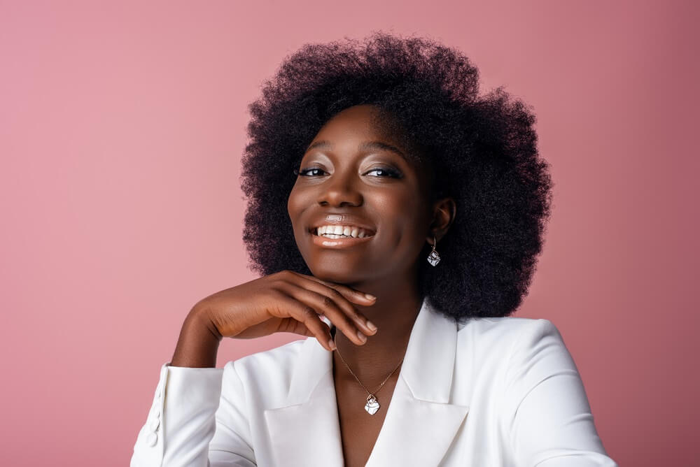 Mulher sorrindo, usando um blaser branco com a mão no queixo em fundo rosa. Imagem ilustrativa do texto sobre franquias investimento baixo.