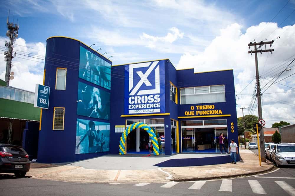 Fachada de uma academia da Cross Experience, com as paredes azuis com detalhes em amarelo, com um grande X branco. Ao fundo, o céu está claro e poucas nuvens brancas. Imagem ilustrativa do texto franquia Cross Experience.