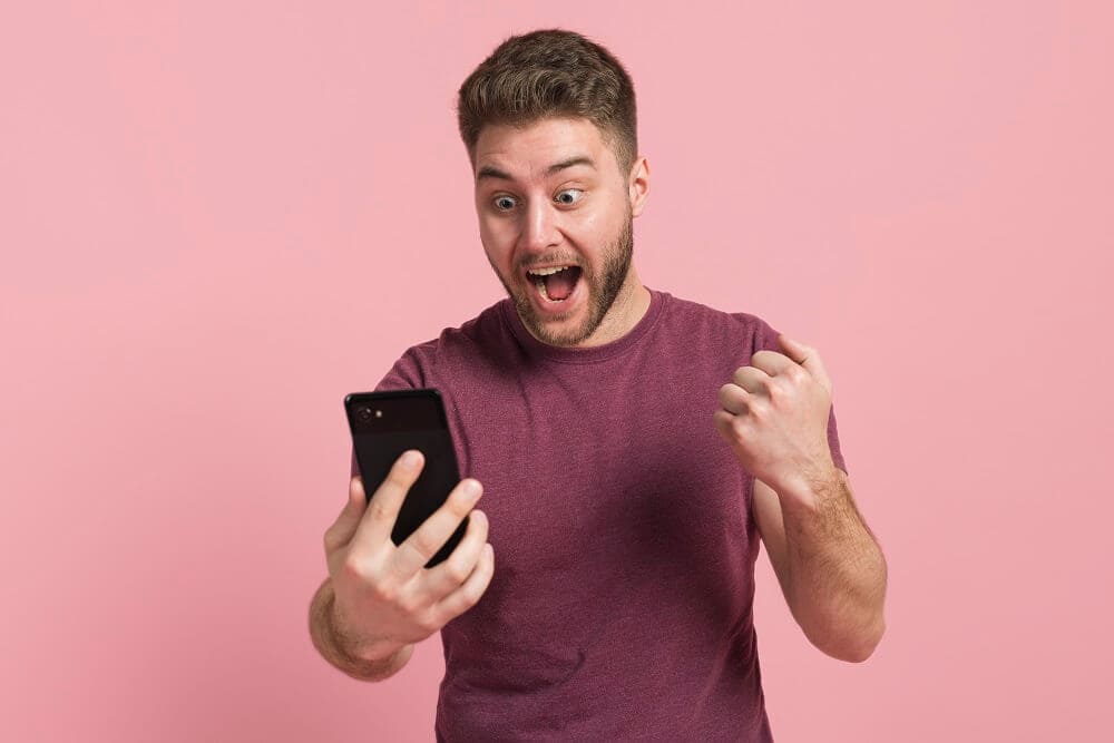 homem de camiseta roxa comemorando enquanto olha o celular ilustraçao de negócios rentáveis com baixo investimento