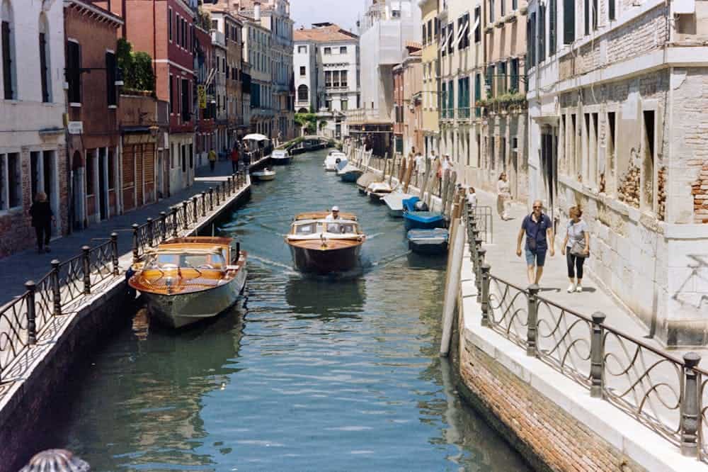 Barcos com detalhes marrons em canal de Veneza. Ilustração do texto sobre agência de viagens física X agência virtual. Qual modelo escolher.