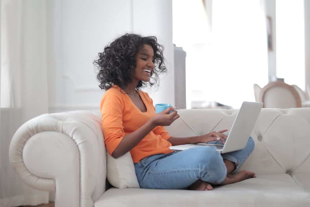 Franquias baratas online 2026 - guia rápido sobre o sistema Mulher sorridente, usando blusa laranja, enquanto trabalha sentada em um sofá, com o notebook no colo. Ilustração do texto sobre franquias para vender online.