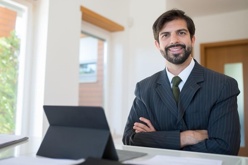 Homem usando um terno cinza escuro sentando em uma mesa escritório sorrindo e com um tablet à sua frente. Imagem ilustrativa do texto investir em franquia vale a pena.