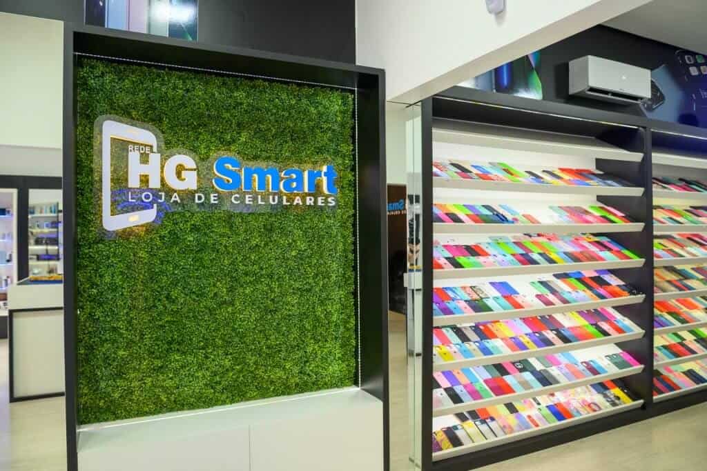 As 128 melhores franquias baratas em 2026 - veja a lista Logomarca da HG Smart na parede. A parede tem folhagem verde e a logomarca tem as cores branca e azul. Ao lado, há uma prateleira com capinhas coloridas para celular.