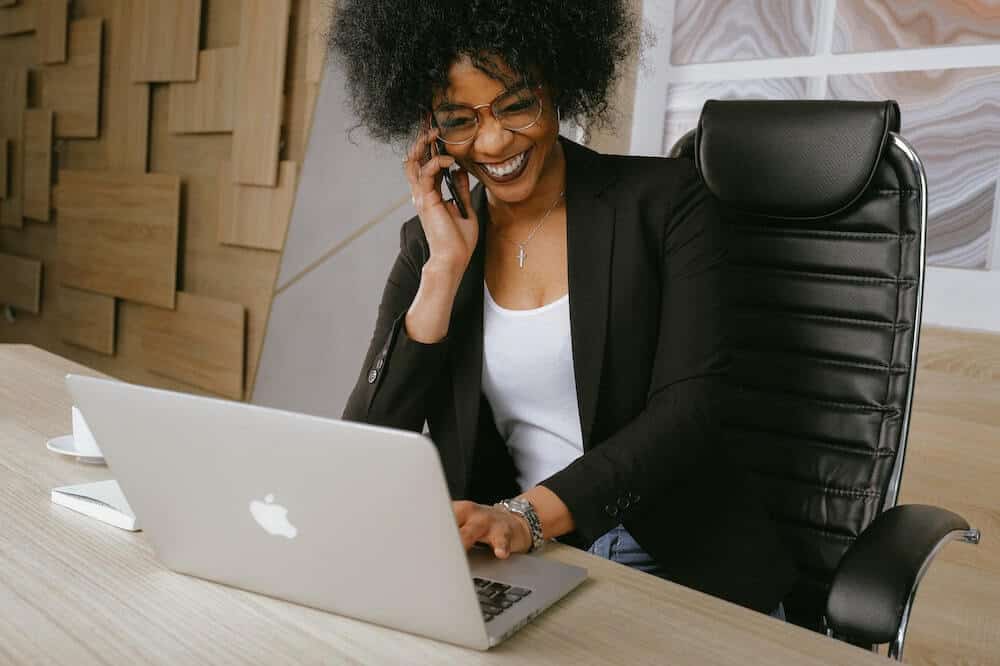 Franquias baratas online 2026 - guia rápido sobre o sistema Mulher sorridentes usando um blase preta, falando ao telefone enquanto analisa um notebook. Ilustração do texto sobre como ser um franqueado online.