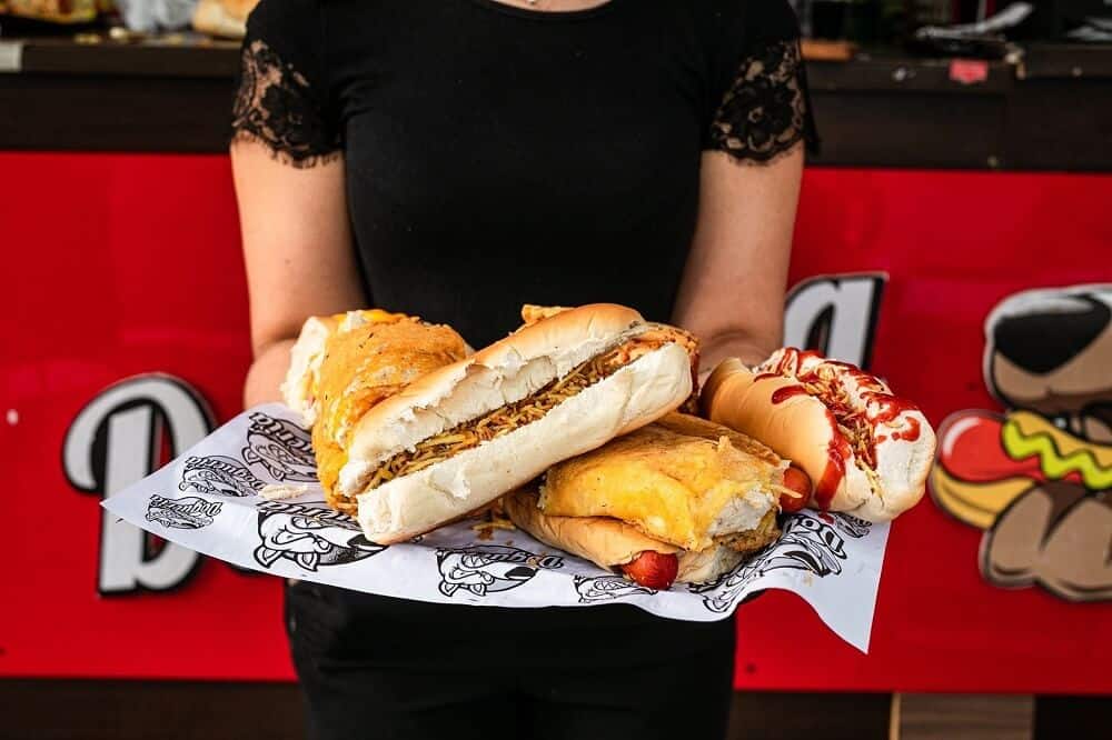 As 128 melhores franquias baratas em 2026 - veja a lista Moça segura bandeja com vários hot dogs, enquanto está em frente a uma logomarca da Dogueria Perdizes