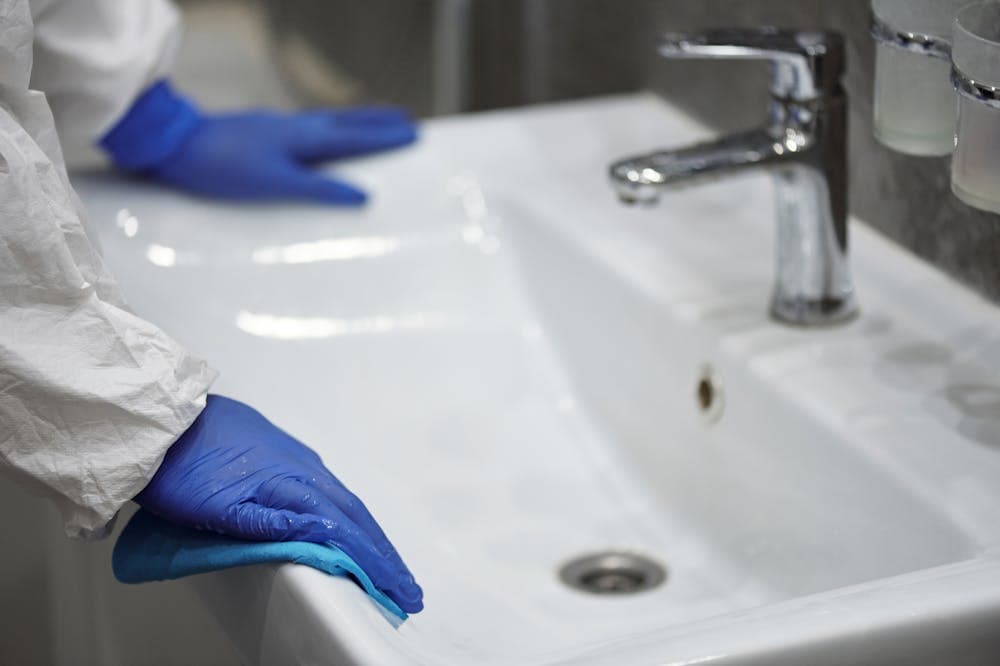 Pessoa usando luva azul, limpando pia de banheiro. Ilustração do texto sobre franquias, empresas, limpeza e conservação.