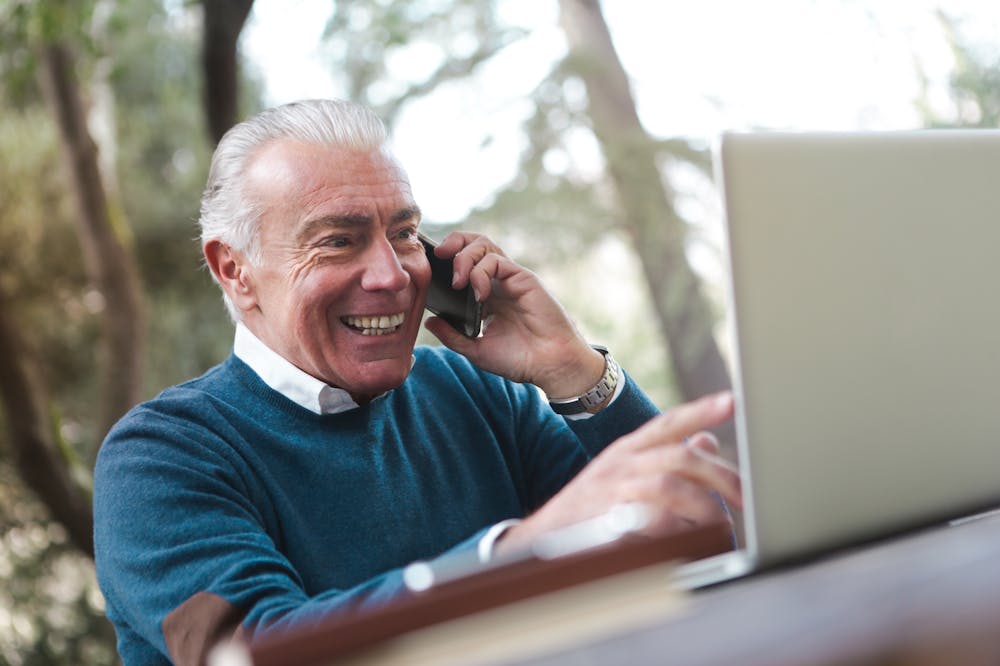 Homem com cabelos brancos, sorridente, sentado em ambiente aberto, enquanto trabalha no computador e fala no celular. Ilustração do texto sobre franqueamento.