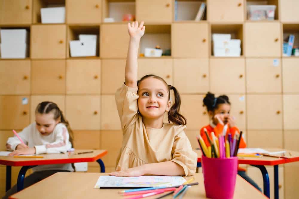 Criança sorridente em uma sala de aula, usando vestido bege enquanto levanta a mão. Ilustração do texto sobre franquia de cursos para crianças.