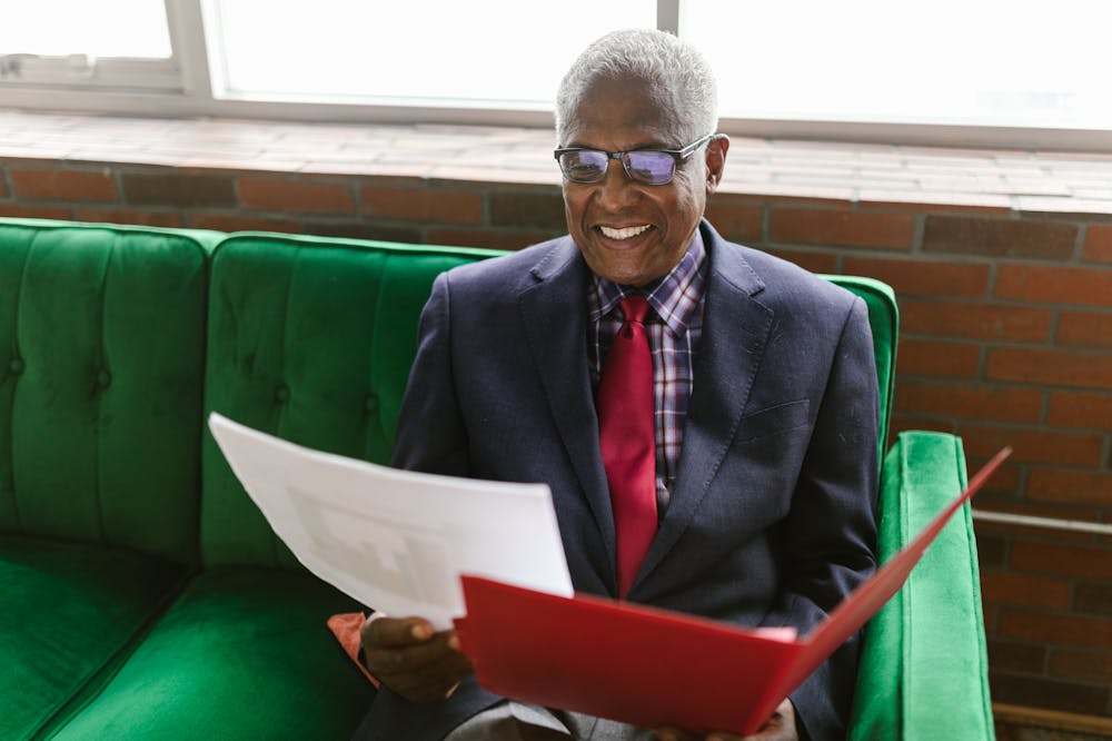 Homem sorridente, usando terno, analisando papéis enquanto está sentado em um sofá verde. Ilustração do texto sobre ser próprio chefe.