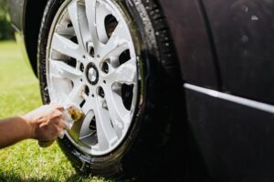 Pneu preto sobre grama verde, enquanto é limpo por uma mão com esponja e espuma. Ilustração do texto sobre franquia de lavagem de carros.