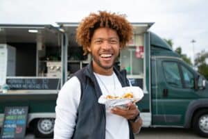 Homem sorridente, usando blusa longa brana e colete cinza, em frente a um food truck, enquanto segura um sanduíche. Ilustração do texto sobre franquias food truck.