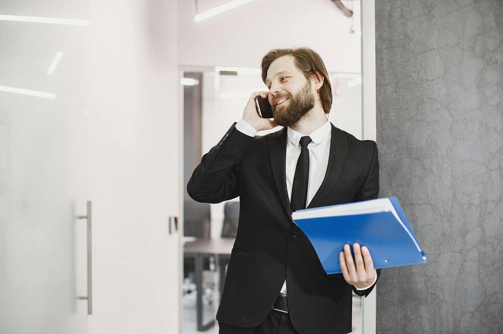 Homem sorridente, usando terno e ao celular, enquanto segura pasta azul. Ilustração do texto sobre franquias para investir com pouco dinheiro.