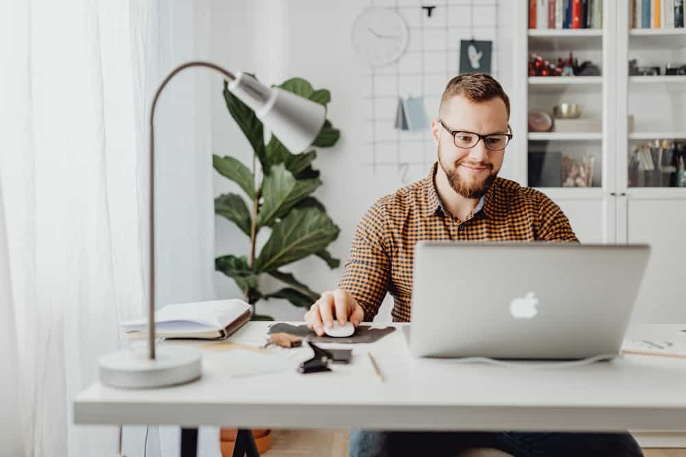 Homem sorridente, usando blusa com manga longa marrom, enquanto trabalha em home office. Ilustração do texto sobre dicas para o seu negócio.