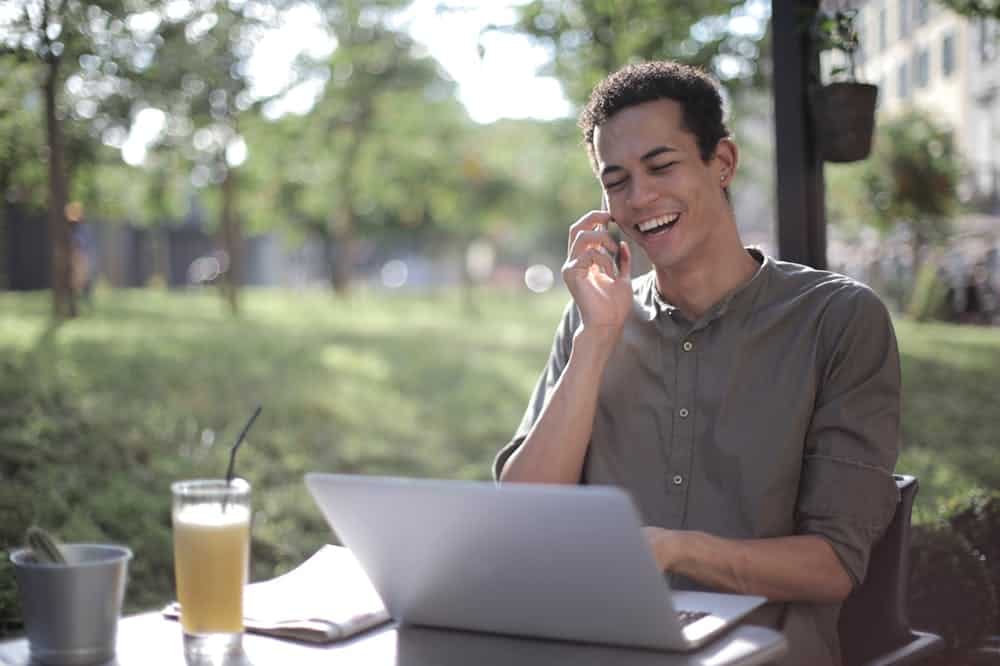 Homem sorridente, sentado em um ambiente arborizado, enquanto trabalha em seu notebook cinza e segura o celular. Ilustração do texto sobre franquias baratas 2024.