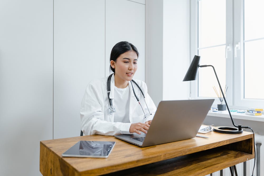 Médica, sorridente, usando jaleco branco, enquanto trabalha em um computador no consultório. Ilustração do texto sobre franquias de saúde.