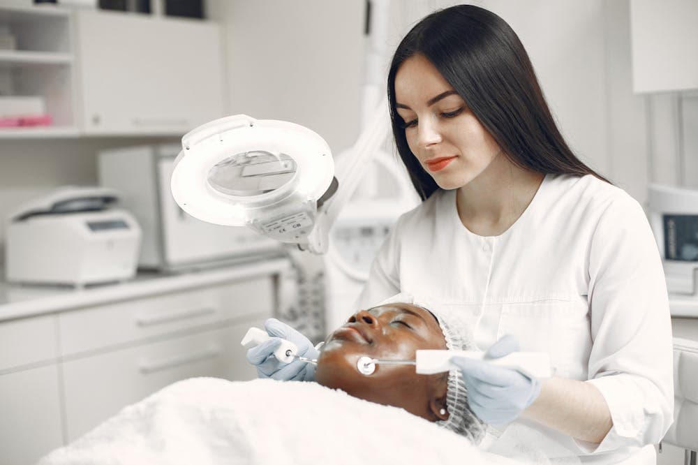 Esteticista concentrada, usando uniforme branco, enquanto cuida da pele da sua cliente. Ilustração do texto sobre melhores segmentos para investir.