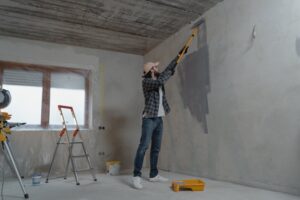Homem atento, usando calça jeans e blusa de flanela, pintando parede com tinta cinza. Ilustração do texto sobre franquia reformas.