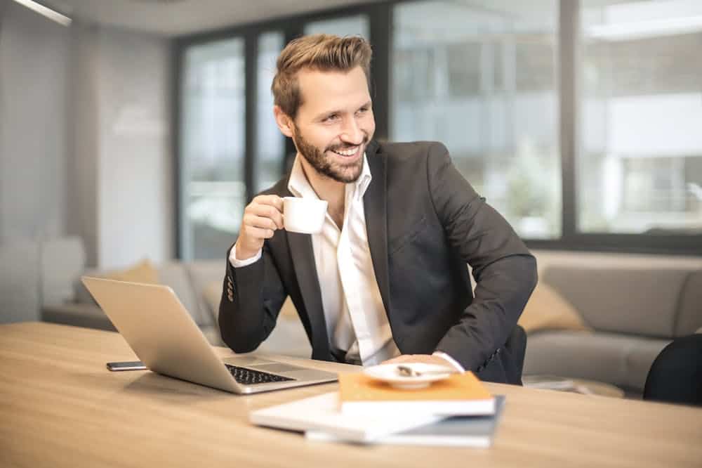 Homem sorridente, em um escritório, trabalhando em frente a um notebook, enquanto segura uma xícara branca. Ilustração do texto sobre características de um empreendedor.