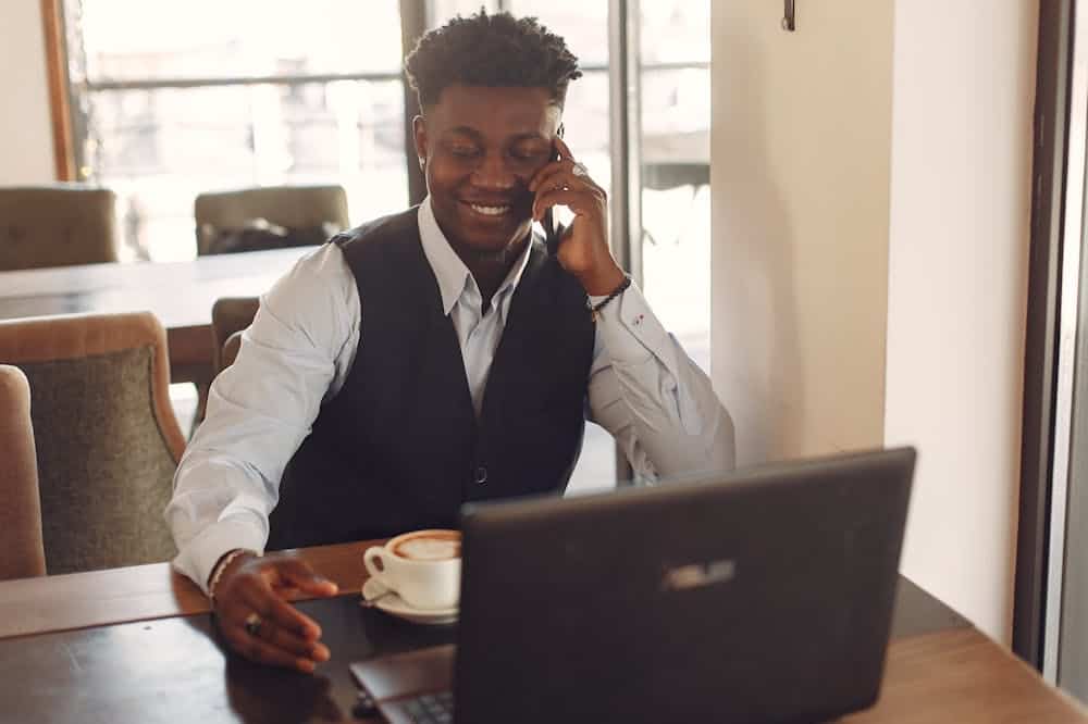 Homem usando blusa social branca e colete preto, sorri ao telefone. Ilustração do texto sobre empreender sendo iniciante.