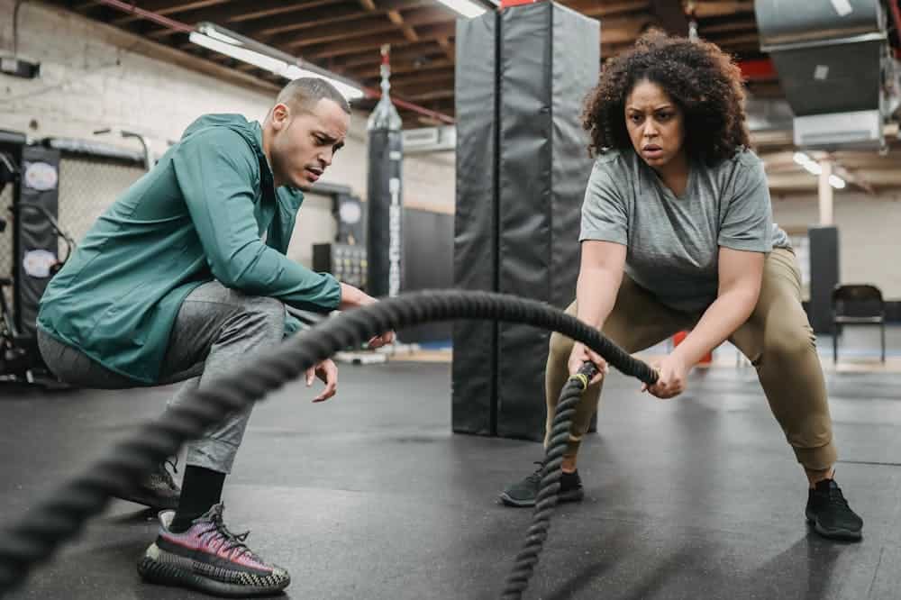 Mulher concentrada, fazendo crossfit, sendo orientada por professor com blusa verde. Ilustração do texto sobre franquia de academia.