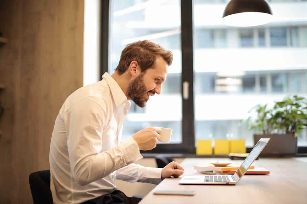 Homem sorridente, usando blusa branca e segurando xícara de café, sentando em frente a um notebook. Ilustração do texto sobre franquias que mais crescem.