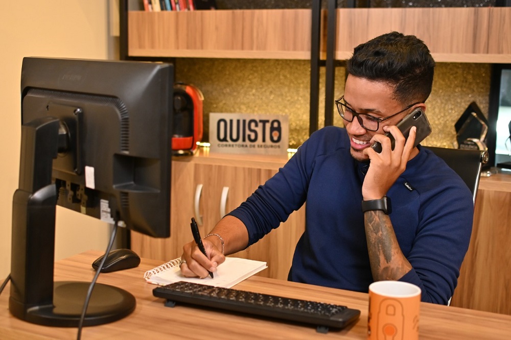 Homem sorridente usando blusa social azul-escuro, enquanto usa o telefone. Ilustração do texto sobre franquia Quisto.