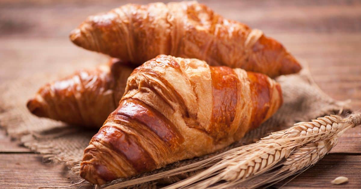 Franquia Croasonho: ganhe dinheiro com croissants artesanais! Croissants frescos e dourados em fundo rústico, símbolo de padaria artesanal e café da manhã delicioso, com trigo como elemento decorativo.