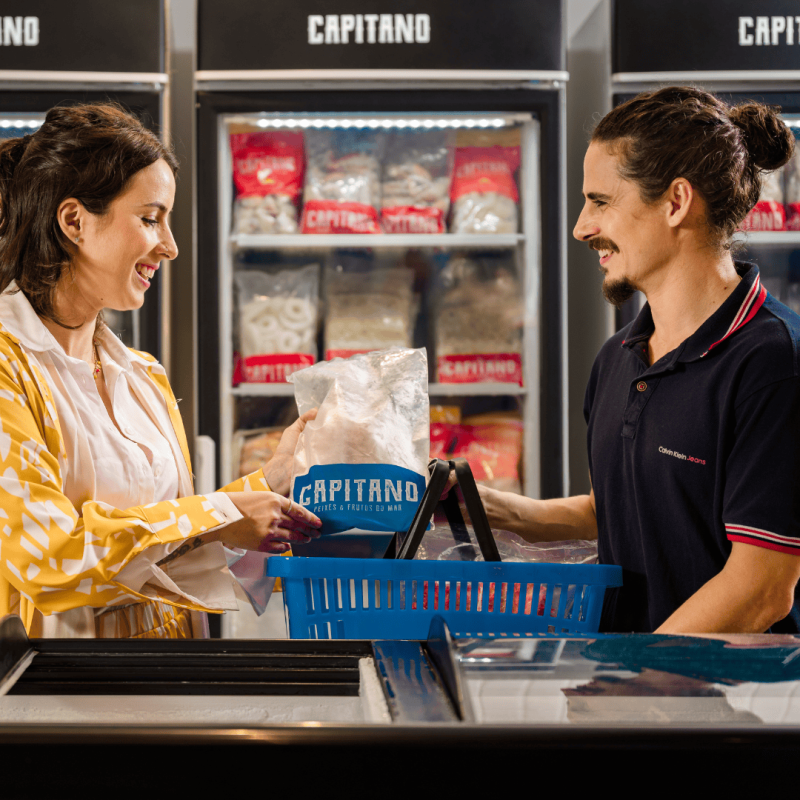 Cliente e atendente com pescada em frente à freezer Capitano.