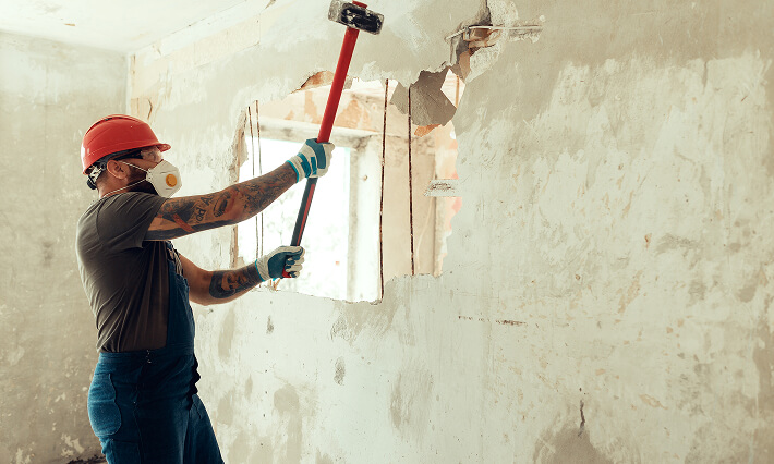 A builder with a hammer in his hands demos an interior wall.