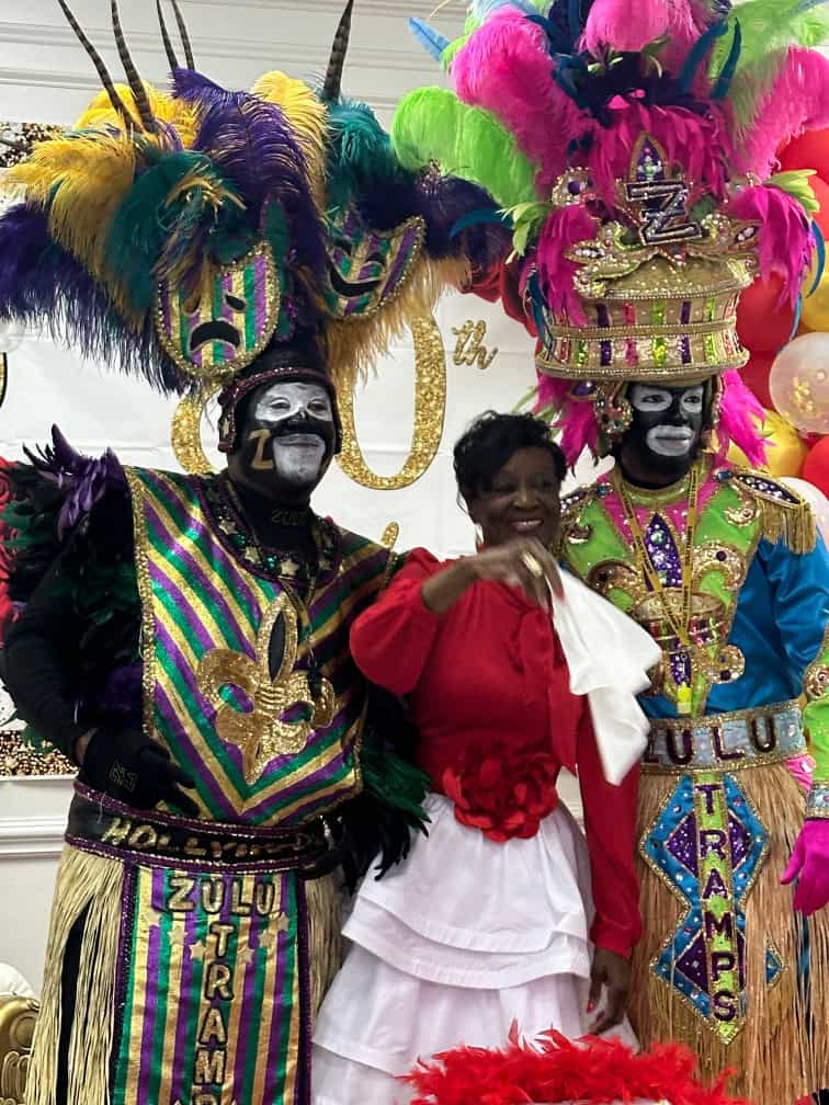 Three people posing for a photo, two in elaborate feathered costumes with face paint, and one wearing a red top with a white skirt, against a decorated background.