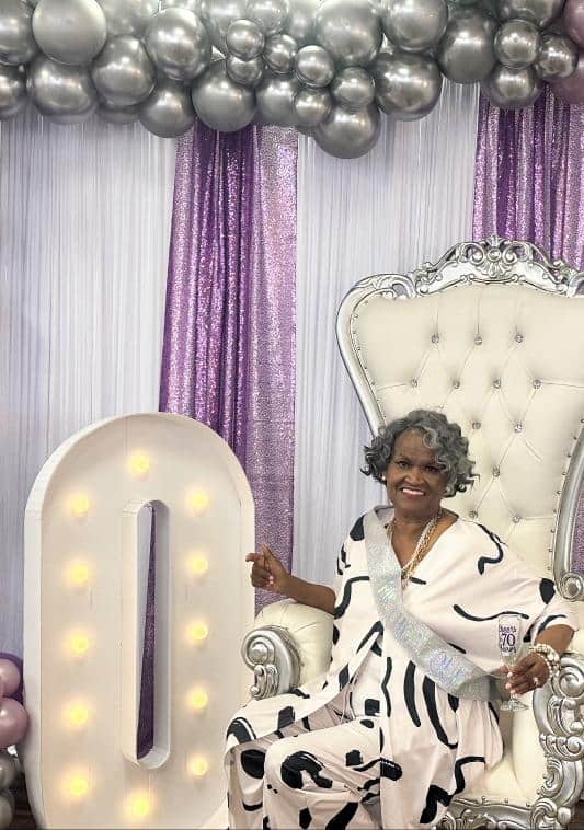 Elderly woman in a sash sits on a white ornate chair next to a lit “0” sign, with silver balloons and purple and white drapes in the background.
