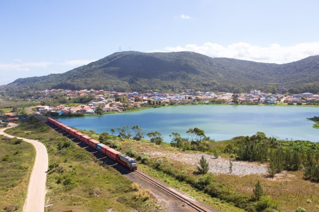 Trem na Ferrovia Tereza Cristina com vista para lago e montanhas ao fundo.