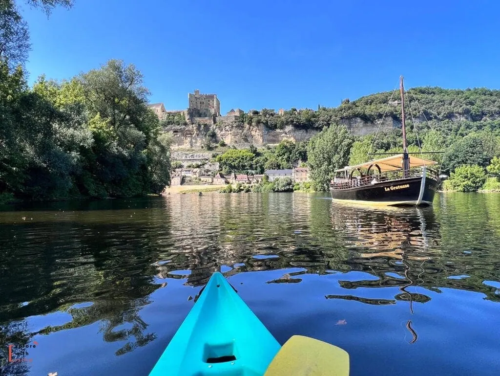 View from a turquoise kayak on the Dordogne River, looking towards Beynac Castle perched high on limestone cliffs. A traditional wooden gabarre boat (La Croisiere) is moored near the riverbank. The medieval village of Beynac-et-Cazenac cascades down the cliff face beneath the castle. The scene is reflected in the calm river water on a bright sunny day, with lush greenery framing both sides of the river.